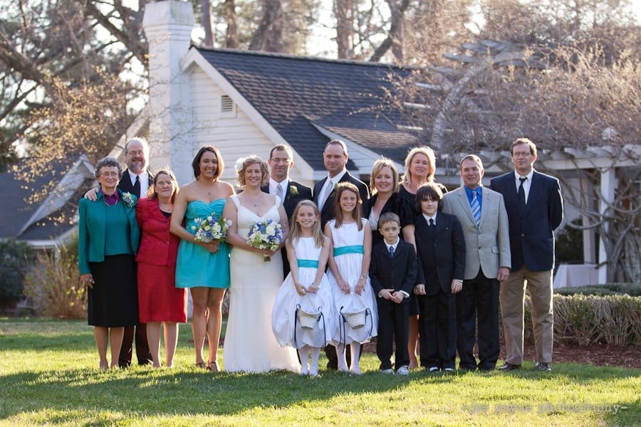 A wedding party poses outdoors in front of a house. The group includes the bride in a white dress, bridesmaids in turquoise dresses, groomsmen in suits, and children in formal wear. The background shows trees and a partially visible house.