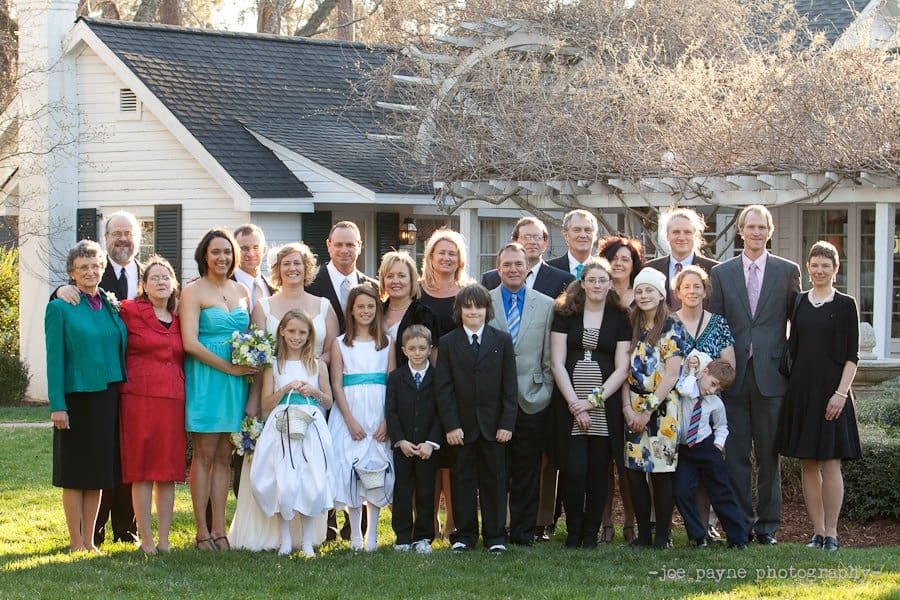 A large group of people, including adults and children, are standing together in front of a white house. Some are dressed formally, suggesting a special occasion. The scene is set outdoors on a sunny day.