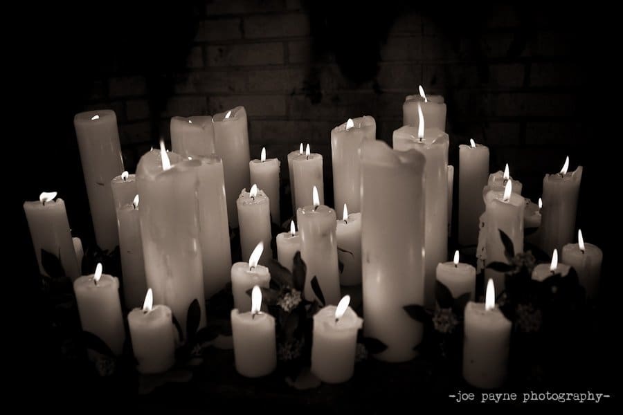 A collection of lit candles arranged closely together, providing a warm and soft glow against a dark background. Some candles have decorative flowers near their bases.