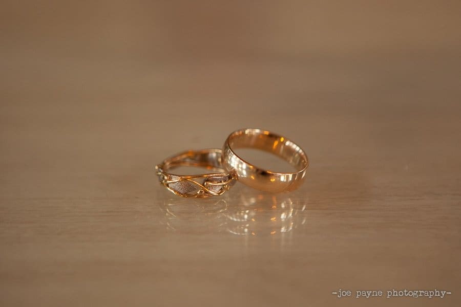 Two gold rings on a reflective wooden surface. One ring is plain and polished, while the other has an intricate, detailed design. Lighting highlights the shine of the metal.