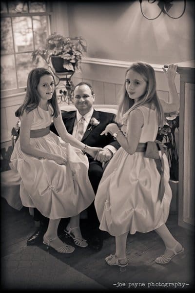 A man sits smiling in a room with two young girls in dresses holding his hands. The girls have long hair and wear matching dresses with sashes. They are inside a room with a window and plants in the background.
