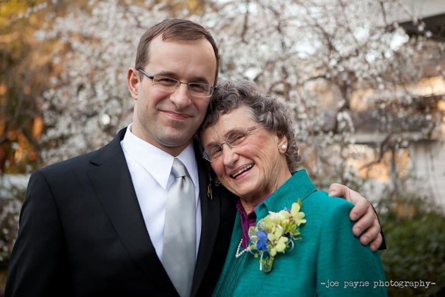 A man in a suit and an older woman with glasses and a teal jacket smile together in front of a flowering tree. The woman has a floral corsage pinned to her jacket.