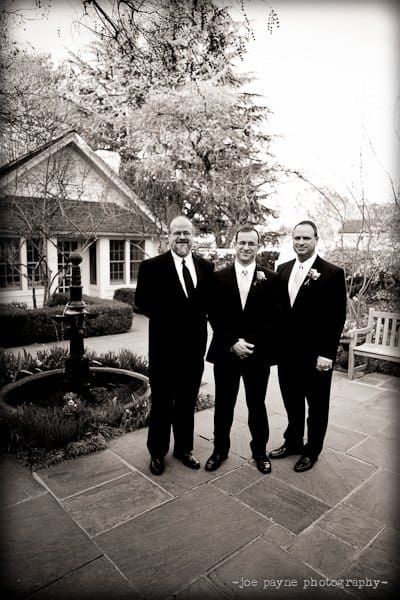 Three men in formal suits stand together outdoors on a stone patio. The setting includes a small building and a fountain. The photo is in black and white.