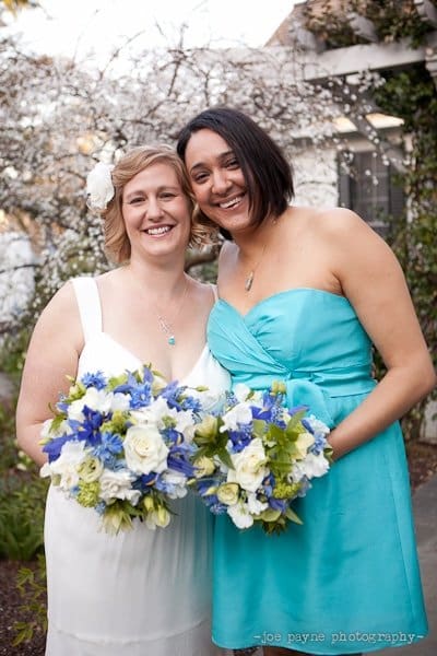 Two women smiling and holding flower bouquets with blue and white flowers. One wears a white dress, the other a turquoise dress. They stand outdoors with blooming trees in the background.