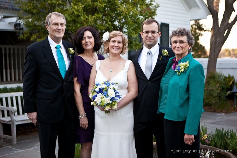A wedding party stands together, smiling. The bride in a white dress holds a bouquet, and the groom wears a black suit and blue tie. They are flanked by two women and a man in formal attire, standing outdoors with greenery in the background.