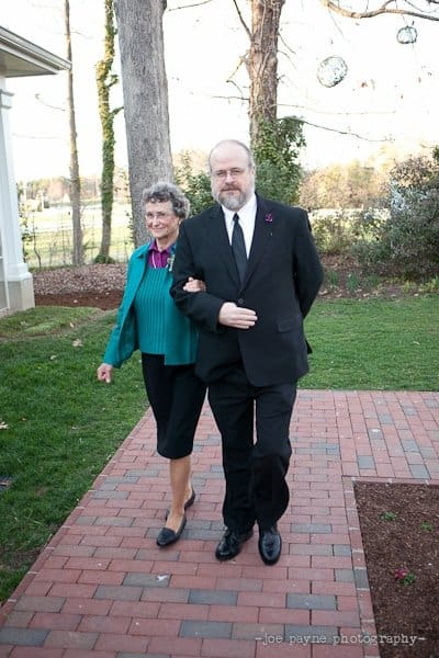 An elderly woman in a blue jacket walks arm in arm with a man in a black suit and tie on a brick path. They are outside near trees, with grass and a building in the background.