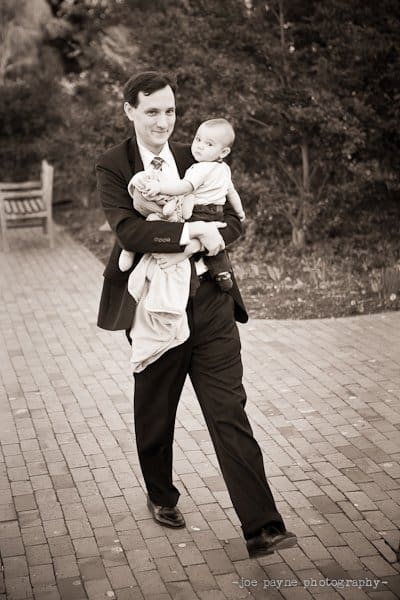 A man in a suit walks on a brick path, carrying a baby and a soft toy. Trees and a wooden bench are in the background. The image is in black and white.
