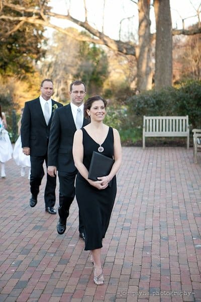 A woman in a black dress walks ahead of two men in suits on a brick pathway, holding a book. They appear to be part of a formal procession. Benches and trees are visible in the background.
