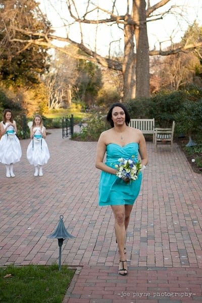 A woman in a turquoise dress walks on a brick path holding a bouquet, with two flower girls in white dresses following behind. The scene is set in a garden with bare trees and green foliage.