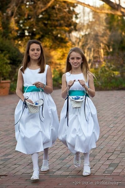 Two young girls dressed in similar white dresses walk side by side on a brick path. They are holding white baskets with blue ribbons. Trees and greenery are visible in the background.