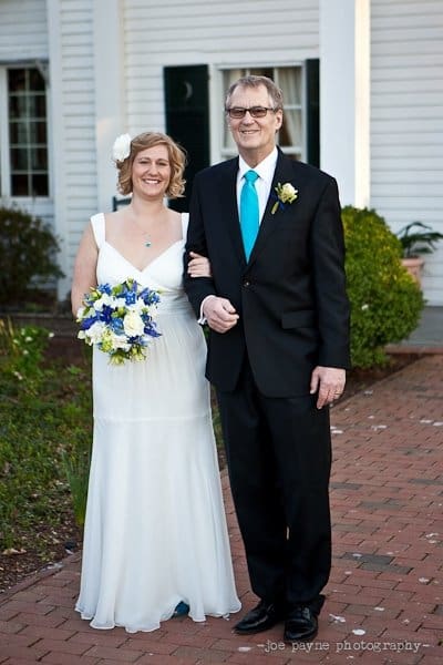 A bride in a white dress and floral headpiece holds a blue and white bouquet, and is walking arm-in-arm with a man in a black suit and turquoise tie. They are outdoors on a brick path with greenery and a white building in the background.