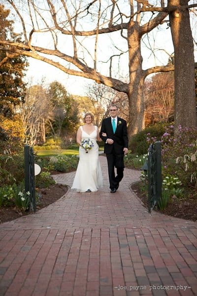 A bride in a white gown holding a bouquet walks arm in arm with a man in a black suit and teal tie. They are on a brick pathway surrounded by greenery and trees under a clear sky.