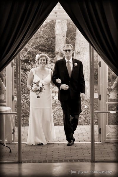 A bride in a white gown with a bouquet walks arm-in-arm with a man in a suit through a doorway, set against a backdrop of trees and outdoor scenery. The image is black and white, and the mood is celebratory.
