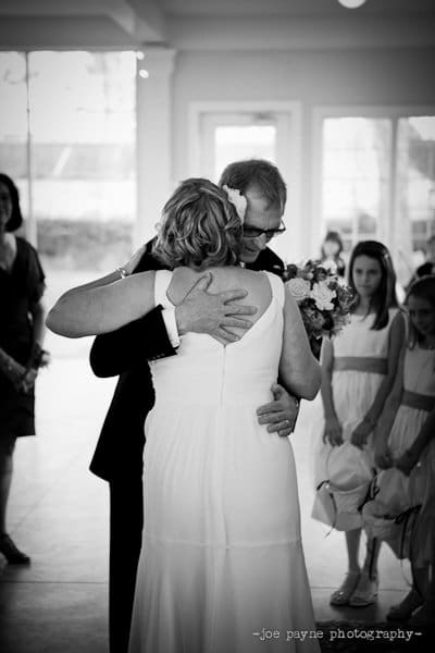 A bride and an older man hug during a wedding. The bride is wearing a long white dress with a flower in her hair, and the man is holding a bouquet. Several people and flower girls in white dresses stand nearby.