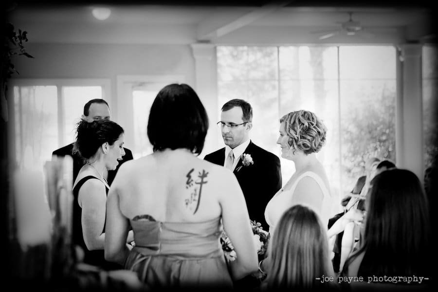 A black and white photo of a wedding ceremony. A bride and groom stand together, surrounded by guests. The officiant and a woman with a tattoo on her back are present. Large windows in the background let in natural light.