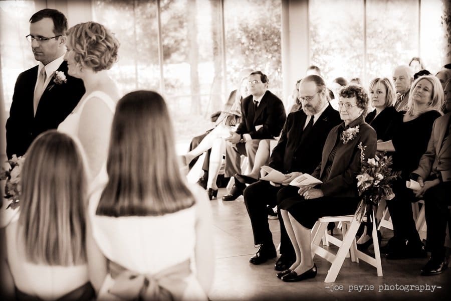 A wedding ceremony with a bride and groom facing each other. Guests sit on white chairs, attentively watching. A man and an older woman in the front row hold a program. Two children stand in the foreground, with long hair and ribbons.