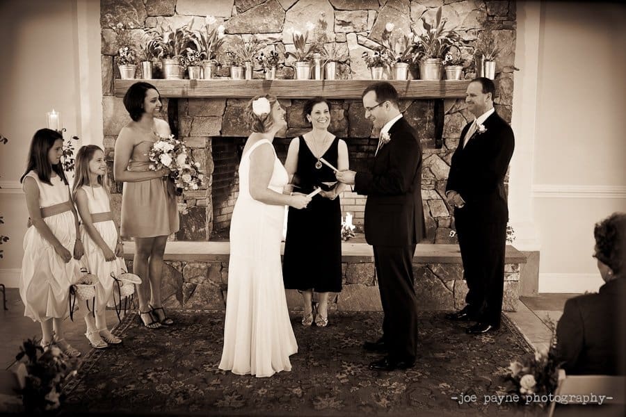 A wedding ceremony with a couple exchanging vows in front of a stone fireplace. The bride wears a white gown and holds the grooms hands. Bridesmaids and a groomsman stand by, along with an officiant. The atmosphere is intimate and elegant.