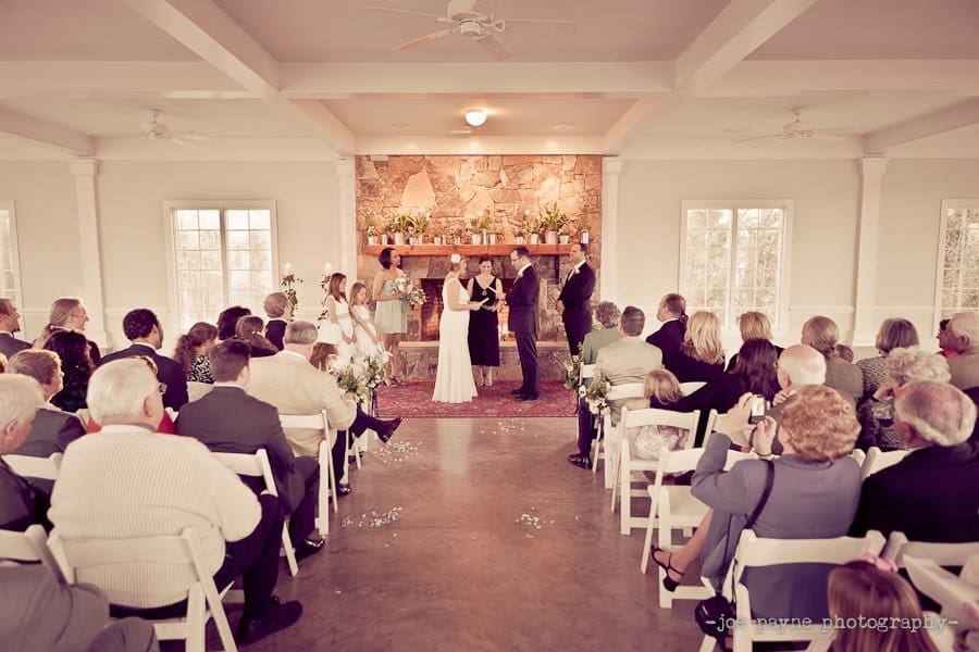 A wedding ceremony in a cozy room with a stone fireplace. The couple stands before an officiant, surrounded by guests seated on white chairs. Bridesmaids and groomsmen stand nearby. The atmosphere is warm and intimate.