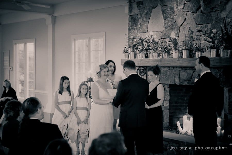 A bride and groom stand before a stone fireplace, exchanging vows during a wedding ceremony. Bridesmaids in white dresses and two groomsmen stand beside them. Guests are seated in the foreground. The setting is elegant and intimate.