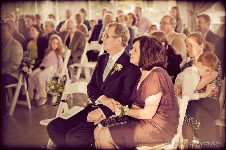 A crowd of people seated at a wedding ceremony. A man and woman sit together in front, holding hands, with flower arrangements nearby. A woman in the back holds a sleeping child. The atmosphere is warm and intimate.