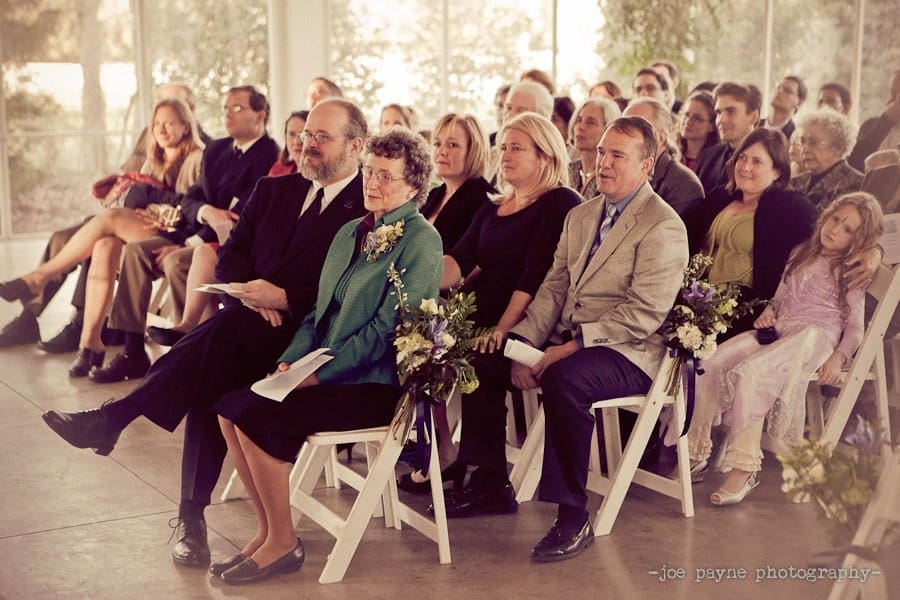 Guests seated at an indoor event, with rows of people on white folding chairs. An elderly couple in the foreground holds floral arrangements, while others in the audience appear engaged and attentive. Soft, warm lighting fills the room.