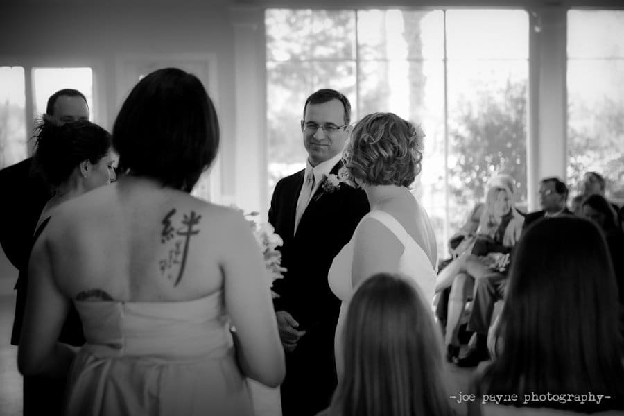 A bride and groom stand facing each other during their wedding ceremony. The brides dress is white and sleeveless. A bridesmaid with a tattoo on her back holds flowers. Guests are seated in the background. The image is in black and white.