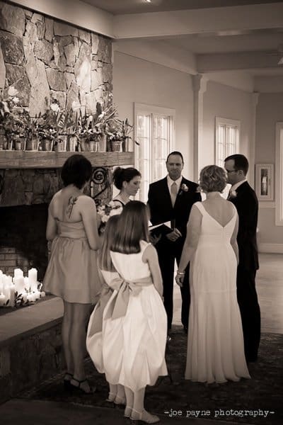 A black and white photo of a wedding ceremony indoors. The couple holds hands, surrounded by three bridesmaids dressed in light-colored dresses. Candles are arranged on a stone fireplace mantel in the background.