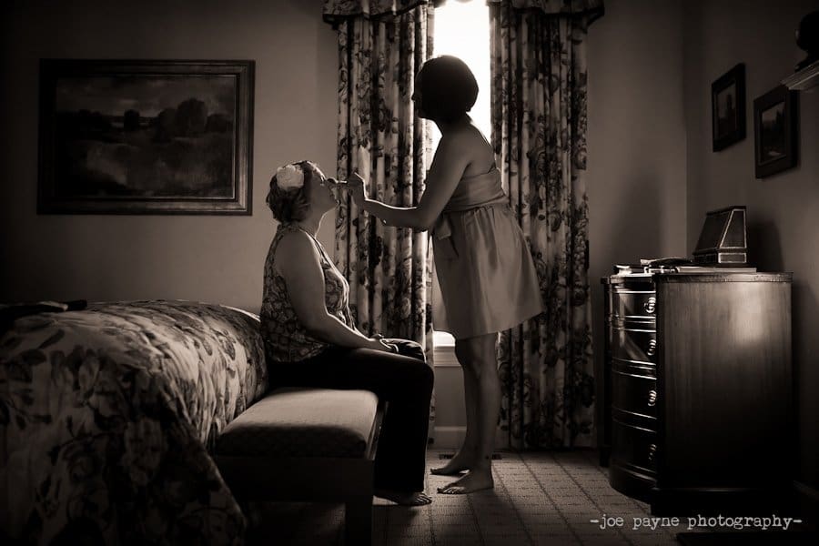 A woman applies makeup to another woman sitting on a bed in a softly lit room. A painting and a dresser are visible. The scene is captured in black and white, creating a warm, nostalgic atmosphere.
