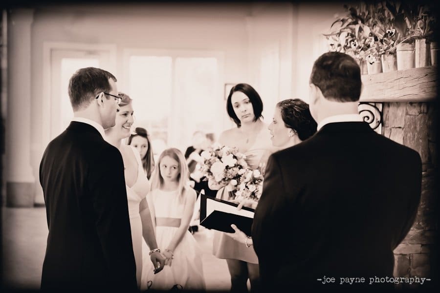 A black and white photo of a wedding ceremony. A bride and groom stand facing each other, smiling. Bridesmaids holding flowers stand nearby. A person is officiating the ceremony. The scene is indoors with soft lighting.