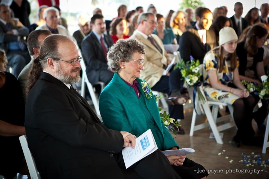 A mature couple sits smiling among a seated crowd at an indoor event. The man wears a suit, and the woman is in a teal blazer with a floral corsage. They hold programs as people around them are engaged, some with flowers.