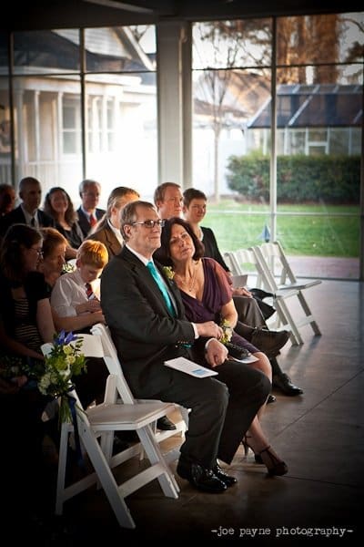 A group of people seated in a light-filled room with large windows. The focal point is a man in a suit and a woman, both sitting in the front row, smiling. Occasional flowers adorn the aisle. The setting appears to be a formal event.