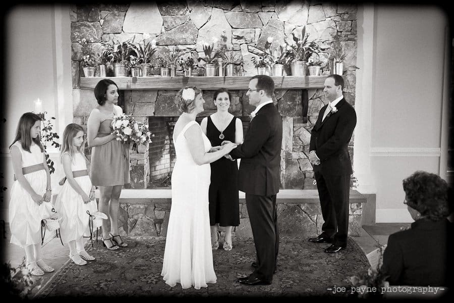 A wedding ceremony with a couple exchanging vows. The bride and groom are standing in front of a stone fireplace. Bridesmaids in dresses and a groomsman in a suit stand nearby. An officiant is present between the couple. Black and white photo.