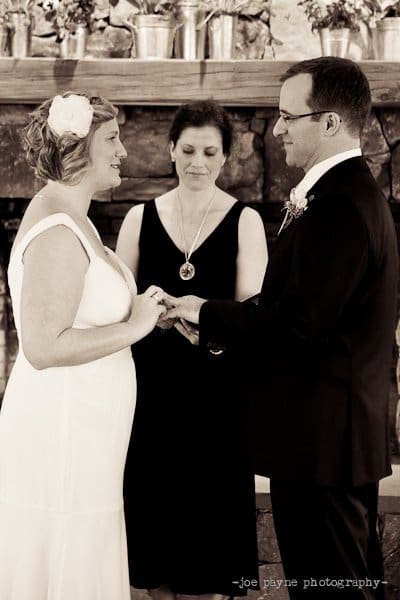 A couple is exchanging vows in a wedding ceremony. The bride is wearing a white dress with a floral hairpiece, and the groom is in a black suit. They are holding hands, with an officiant standing between them. The setting is indoors with a stone backdrop.