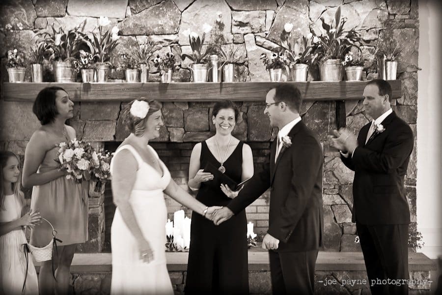 A joyful couple holds hands during their wedding ceremony in front of a stone fireplace. Three attendants stand nearby, one holding a bouquet and another applauding. Everyone is smiling, creating a warm and celebratory atmosphere.