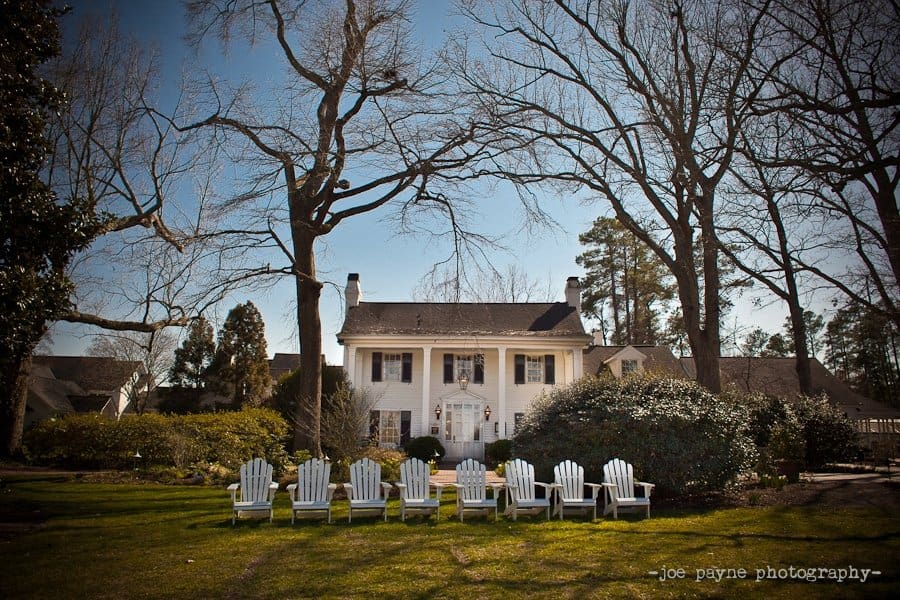 A large, white two-story house with a symmetrical facade and dark shutters stands amidst tall, leafless trees. In the foreground, several white Adirondack chairs are neatly arranged on a green lawn. The sky is clear and sunny.