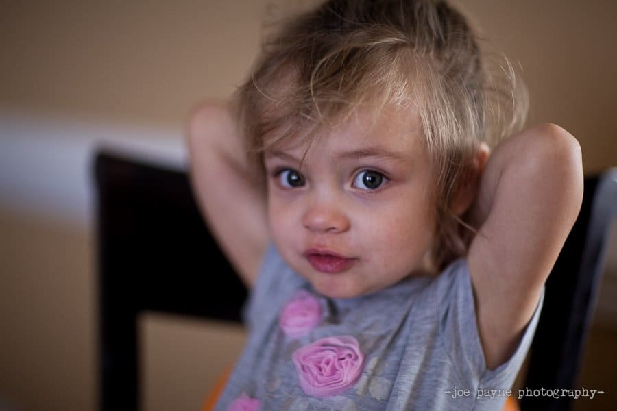 A young child with tousled hair and big blue eyes sits with both hands behind their head, wearing a gray shirt decorated with pink fabric roses—a peaceful pause in the midst of spring cleaning. The background is softly blurred.