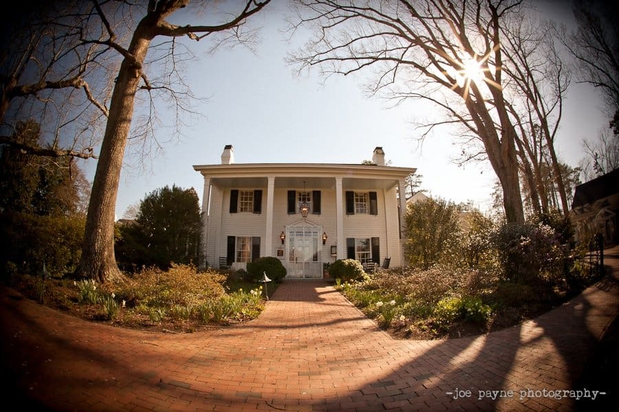 A large two-story white colonial-style house is surrounded by tall trees under a clear blue sky. The sun peeks through the branches, and a brick path leads to the entrance, flanked by shrubs and blooming flowers.