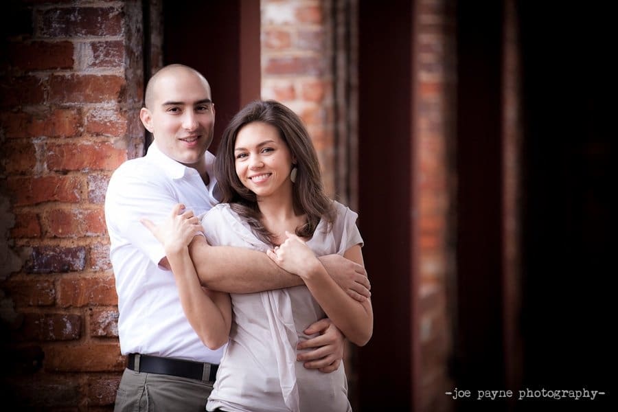 A couple stands smiling and embracing in front of a brick wall during their Greenville engagement session. The man, in a white shirt, stands behind the woman with long brown hair; both look at the camera, appearing happy and relaxed.