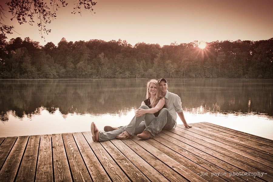 A couple sits on a wooden dock by a calm lake at sunset, smiling and leaning against each other with trees and the sun in the background—capturing the perfect moment for a Raleigh engagement shoot.