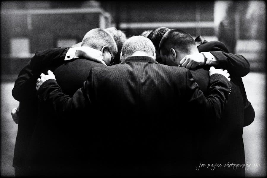 A group of men in suits stand in a close huddle with their arms around each other’s shoulders, heads bowed together in a moment of unity or prayer. Captured by a Raleigh wedding photographer, the black and white image radiates timeless camaraderie.