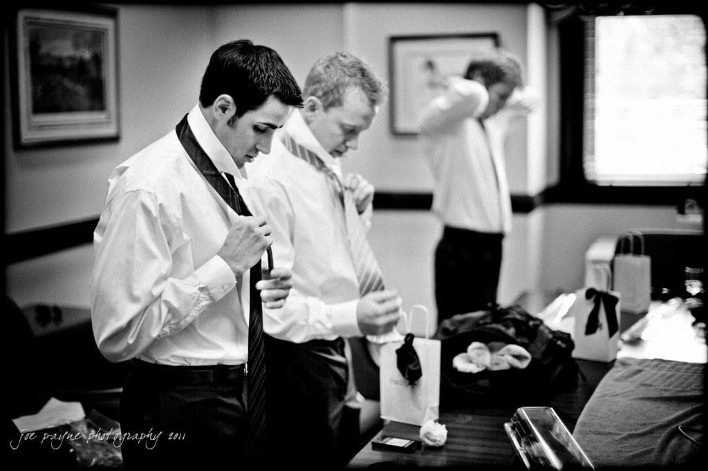 A black-and-white photo of three men in a room getting ready, adjusting their ties. Gift bags with tissue paper are on the table. The room has framed pictures on the walls and a window with blinds.