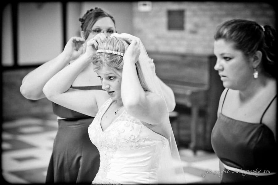 raleigh wedding photographer - jenna and ryan A black and white photo of a bride adjusting her veil with the help of two bridesmaids. The bride wears a beaded dress and tiara. The bridesmaids are in matching dresses. They are indoors, possibly in a church or reception hall.