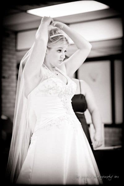 raleigh wedding photographer - jenna and ryan A bride in a white gown adjusts her veil with both hands. The photo is in black and white, and she stands indoors with a blurred background. A person in a darker outfit is partly visible behind her.