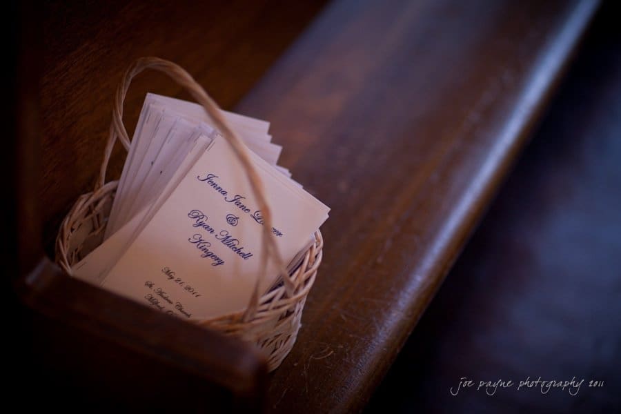 raleigh wedding photographer - jenna and ryan A wicker basket holds several folded papers, likely event programs, on a wooden bench. The text on the programs appears to be formal, possibly for a wedding or ceremony. The lighting is soft, creating a warm atmosphere.