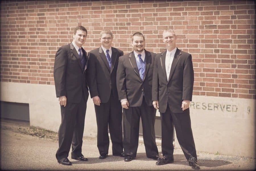 raleigh wedding photographer - jenna and ryan Four men in suits stand side by side in front of a brick wall. They are smiling and appear to be posing for a formal photo.