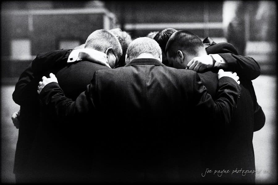 raleigh wedding photographer - jenna and ryan A group of men in formal suits huddle together, arms around each other, in a moment of unity and camaraderie. The black and white image captures the backs of their heads in a dimly lit setting.