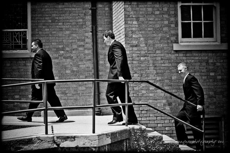 raleigh wedding photographer - jenna and ryan Three men in suits walk along a ramp next to a brick building. They appear to be moving in a line, one after the other, under a rail. The scene is in black and white, with a slightly angled perspective.