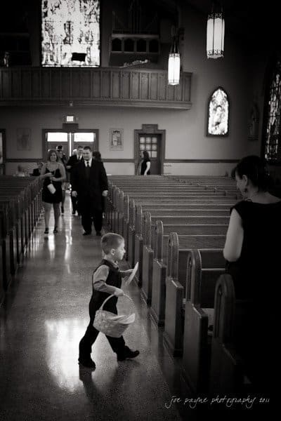 raleigh wedding photographer - jenna and ryan A black and white photo of a young boy running down a church aisle carrying a basket, possibly as a ring bearer or flower boy. Several adults walk behind him, and stained glass windows can be seen in the background.