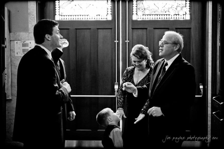 raleigh wedding photographer - jenna and ryan A black and white photo of three adults and a child in formal attire. They stand near wooden double doors, engaging in conversation, with smiles and an atmosphere of celebration. The child looks up at one of the adults.