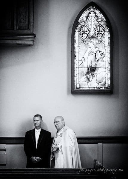 raleigh wedding photographer - jenna and ryan A black and white photo of two men standing in a church. One is wearing a suit, and the other is in religious robes. A stained glass window depicting a religious figure is visible above them.